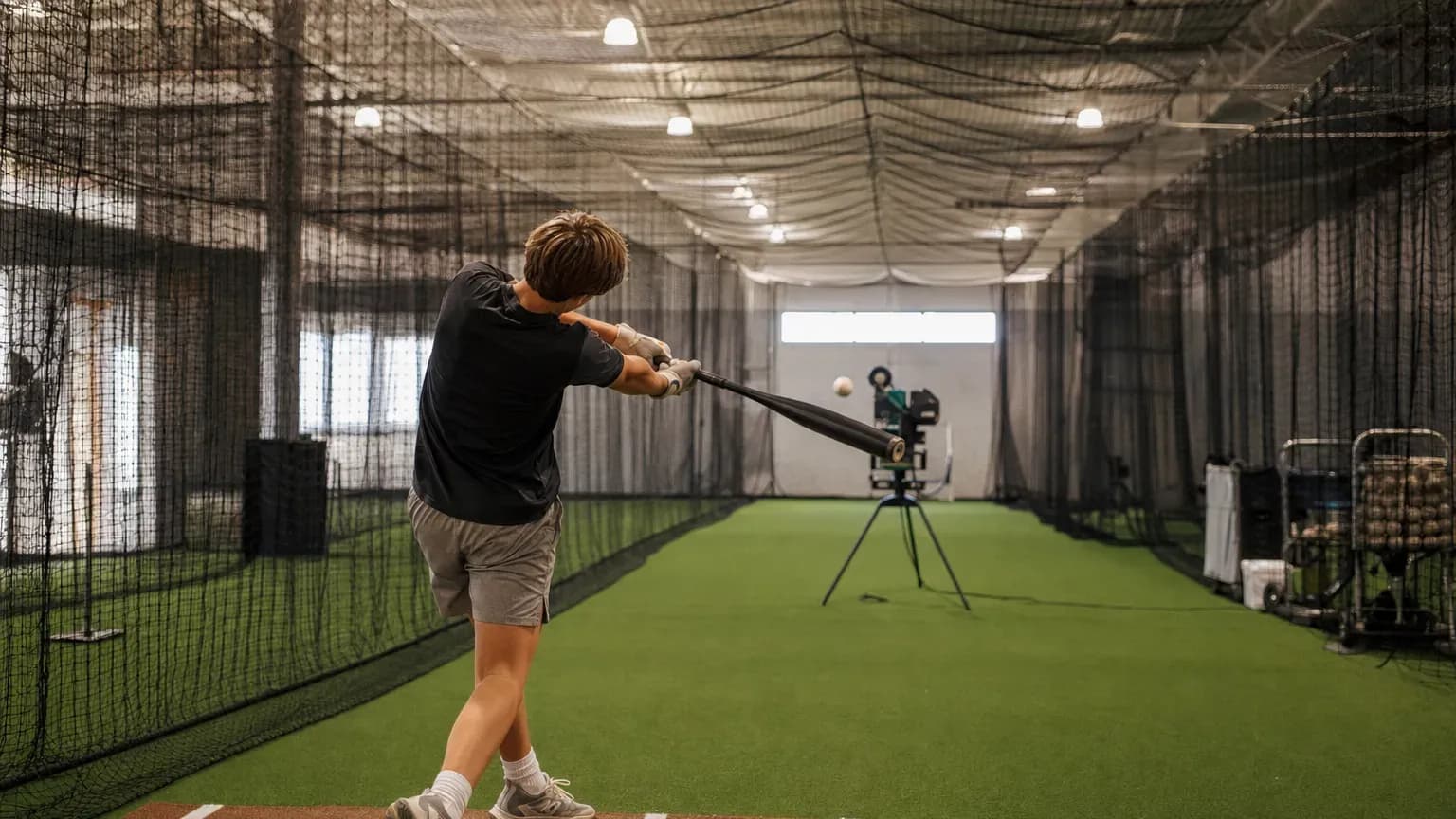 Indoor batting cage tunnels with netting and a player hitting against a pitching machine.